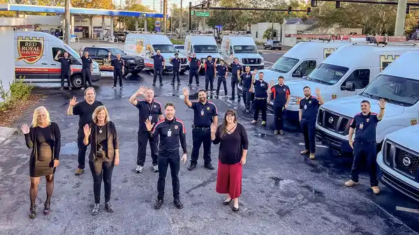 A group of people in uniforms and business attire stand in a parking lot, waving at the camera, with multiple company vans parked behind them—capturing the spirit of our team in this authentic about us moment.